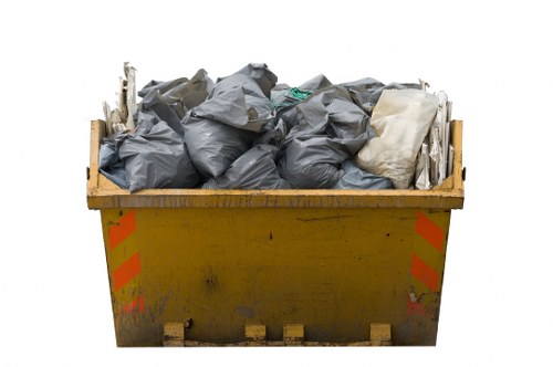 A large yellow industrial skip filled with a mixture of black, grey, and white garbage bags and assorted debris, located outdoors on a paved surface. The skip has red reflective safety markings on its sides, appears weathered with some dirt and scuff marks, and is positioned in an open area. The image shows the debris piled high above the edges of the skip, indicating it is nearly full. The background is plain white, with no additional environmental details visible. This scene is representative of rubbish removal activities undertaken by companies such as Office Clearance Mill Hill in the Middlesex area, supporting local rubbish clearance services in Mill Hill and surrounding locations.