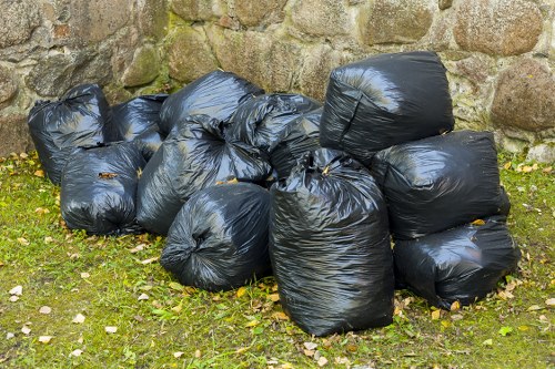 A collection of multiple black plastic rubbish bags, filled and knotted at the top, are gathered on a grassy outdoor area in front of a stone wall. The bags are irregularly shaped, with some appearing tightly packed and others slightly bulging, indicating a variety of waste contents. The surface of the bags is shiny and smooth, reflecting light, and they are positioned closely together, with a few overlapping. The ground beneath them is covered in grass with scattered leaves and small stones, typical of an outdoor environment in a residential or garden setting in Mill Hill. The stone wall in the background provides a natural and rustic backdrop for the scene, suggesting proximity to a property exterior where rubbish collection or clearance services might be carried out by Office Clearance Mill Hill as part of their rubbish removal offerings.