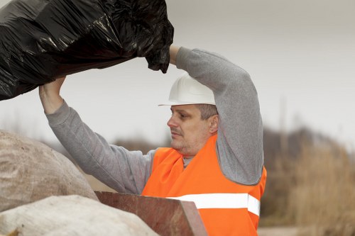 A man wearing a white safety helmet and an orange high-visibility vest is outdoors, lifting a large black trash bag filled with rubbish above his head, preparing to dispose of it into an unknown container or skip. The background shows an overcast sky and a blurred natural landscape with dry grass and sparse trees, indicating an outdoor setting likely related to waste collection or clearance work in the Mill Hill area. His clothing and the presence of large waste bags suggest involvement in rubbish removal or site clearance activities, consistent with services offered by Office Clearance Mill Hill. The scene emphasizes the manual effort involved in rubbish collection, with the man focused on the task at hand, highlighting the physical nature of waste management tasks carried out in the local area.