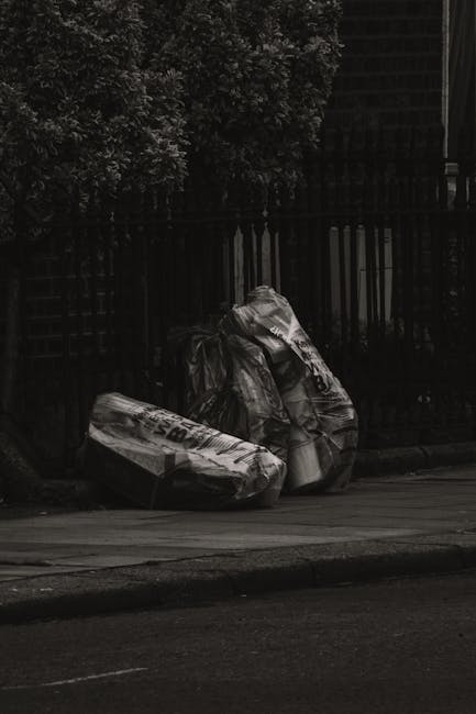 A collection of overflowing rubbish bins and scattered waste bags situated on a paved sidewalk in front of a commercial area. The foreground features a silver and a black bin, both filled with mixed p