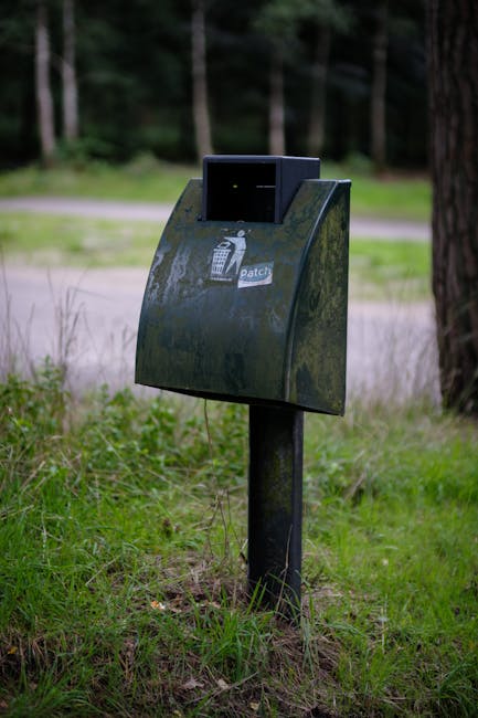 A black, weathered litter bin with a rounded front and a sloped top, mounted on a metal post embedded in grassy ground. The bin features a small, rectangular opening at the top for rubbish disposal an