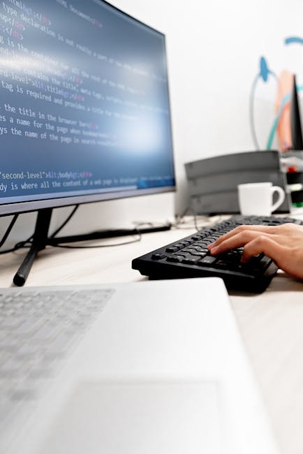 A close-up view of a silver laptop with a black keyboard, positioned on a light wooden desk. The screen displays lines of colorful code on a dark background, indicating programming or software develop