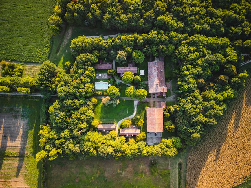 A large historic red-brick building with multiple tall chimneys and a traditional tiled roof, situated amidst a well-maintained garden with lush green grass, various shrubs, and mature trees. The buil
