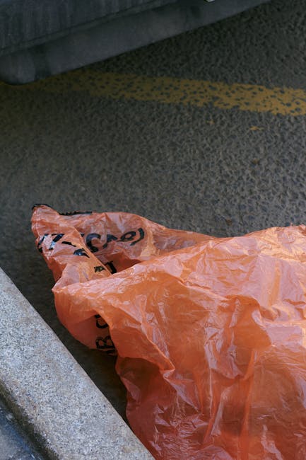 A person with long red hair, wearing a white shirt and a bright orange sleeveless cardigan, is holding an open brown paper bag filled with waste, possibly paper or lightweight rubbish, in a room with