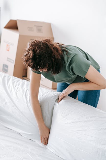 A person with curly brown hair, wearing a green t-shirt and blue jeans, is in a room with boxes in the background, actively removing or unwrapping a large white mattress or fabric item. The individual